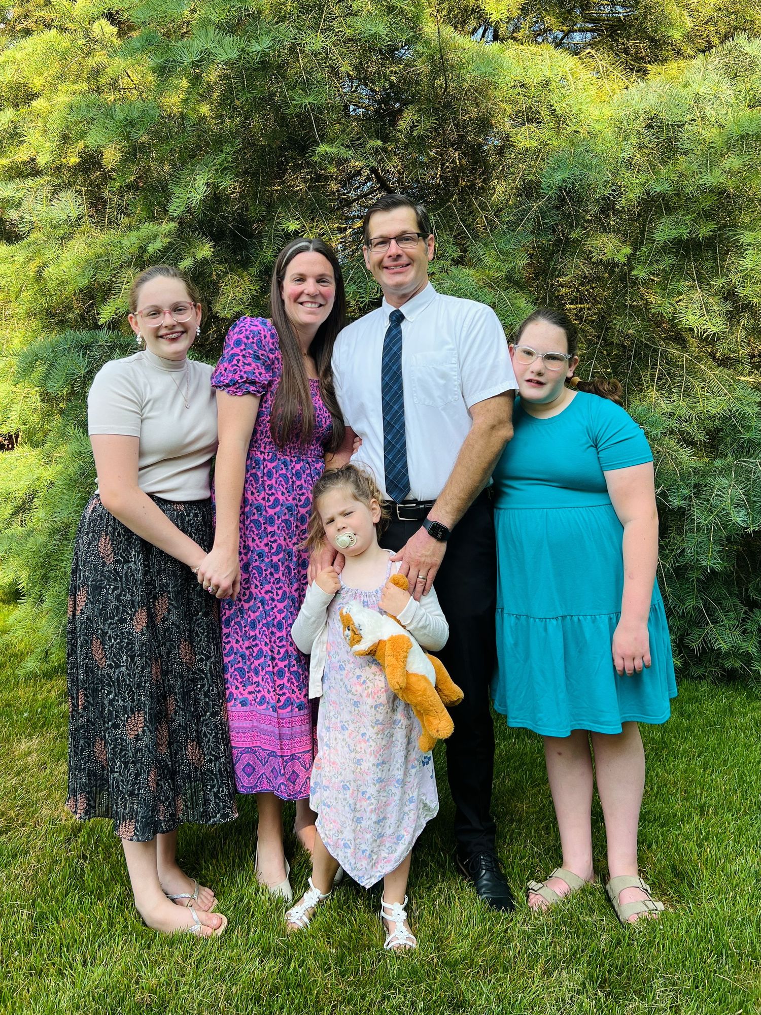 Family of five poses outdoors near evergreen trees with father in white shirt and tie.