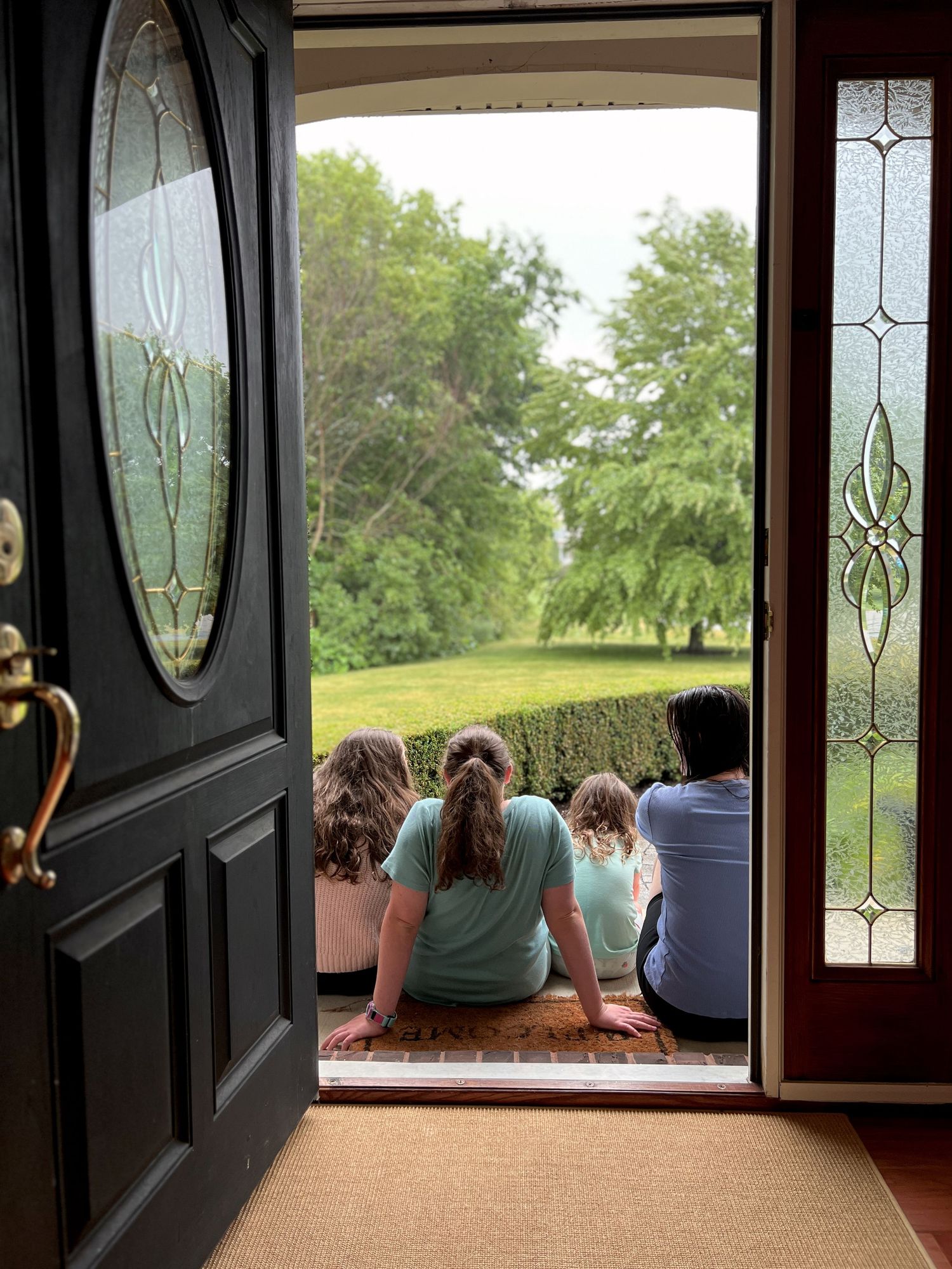 Children and mom sitting in open doorway watching green yard on cloudy day.