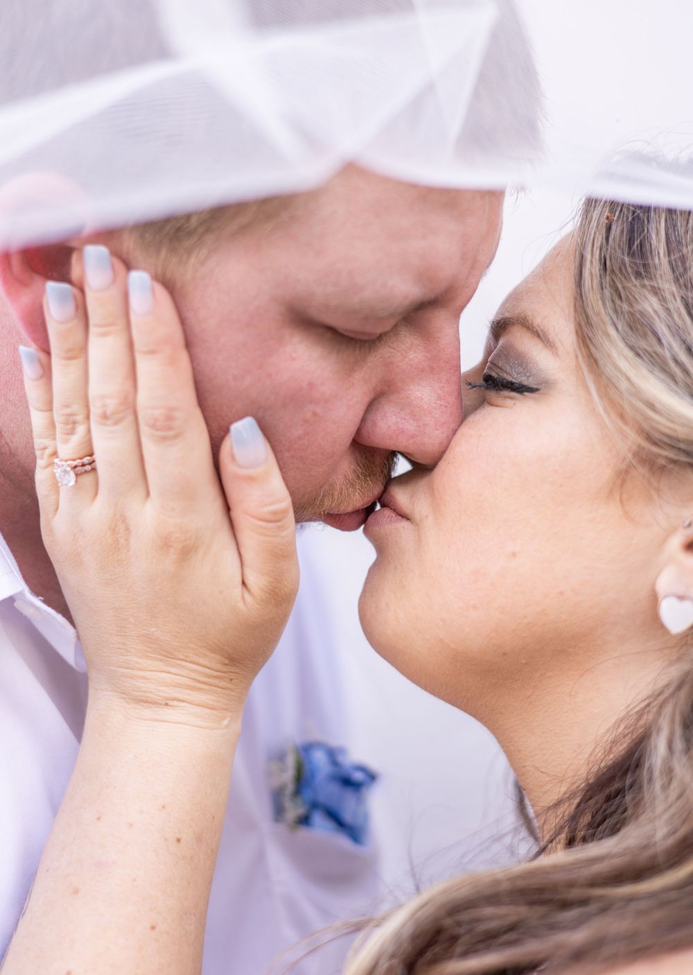 A tender intimate moment shared under a white veil as hands softly touch faces while kissing.