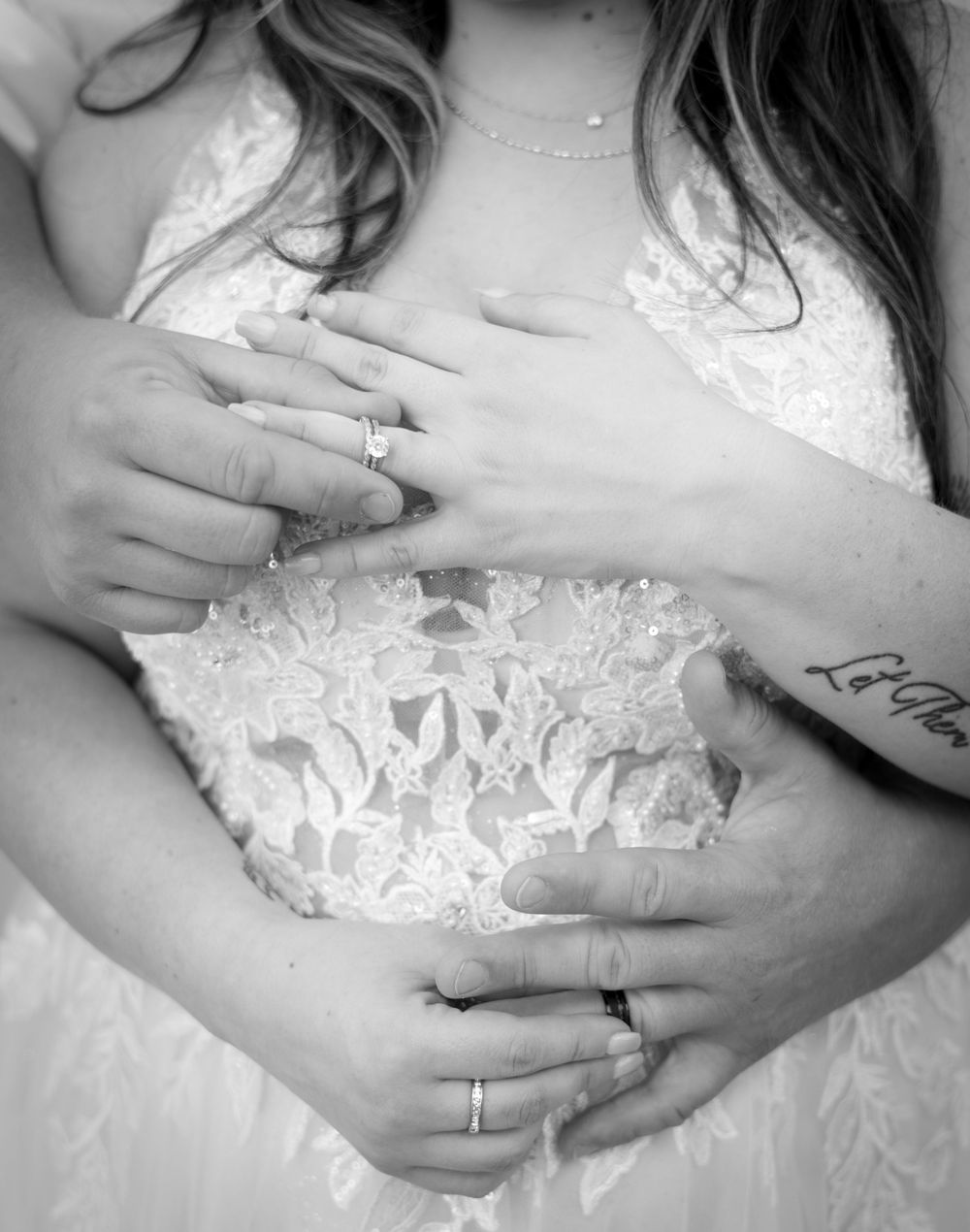 Black and white close up shot of hands with rings embracing a white lace wedding dress.
