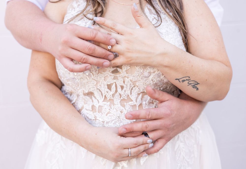 Close-up of hands with rings embracing a white lace wedding dress bodice with a small visible arm tattoo.