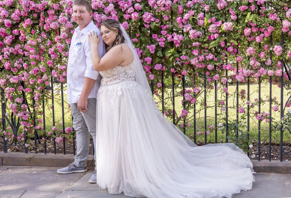 A happy couple poses in wedding attire against a beautiful backdrop of pink roses and an iron fence.