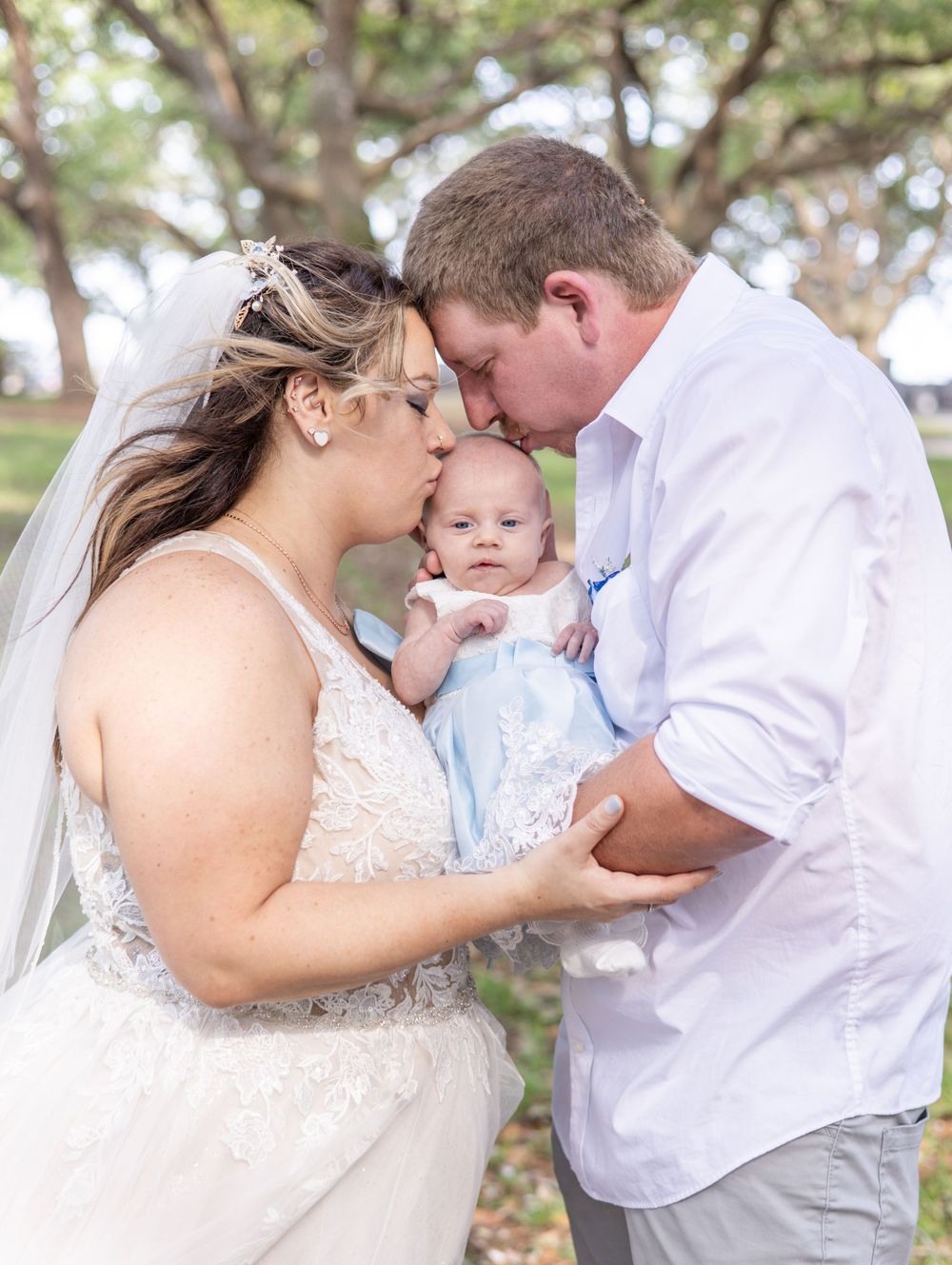 A tender family moment outdoors with a small baby being held and kissed by parents in wedding attire.