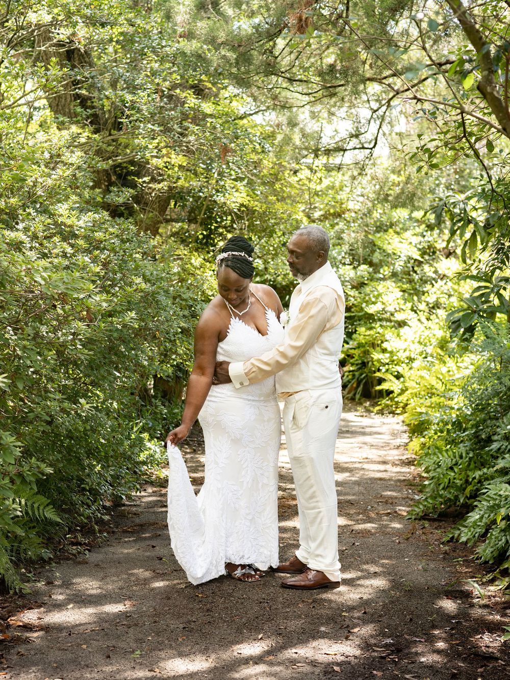 A couple shares a romantic moment on a sunlit forest path while wearing elegant white wedding attire.