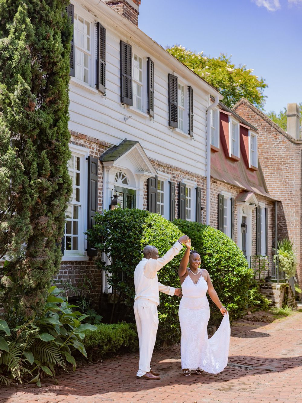 A couple in white attire dancing together on a brick path in front of a charming colonial style house with black shutters.