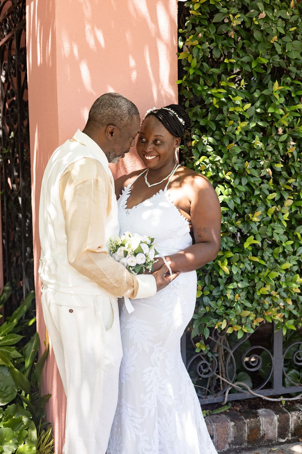 A joyful couple in white attire share an intimate moment against a pink wall and green foliage backdrop.
