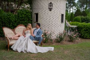A couple shares tender moments in front of a white brick wall surrounded by lush greenery at an outdoor venue.