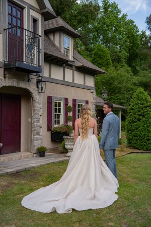 A couple in wedding attire stands together on a lawn in front of a Tudor-style house with stone accents.