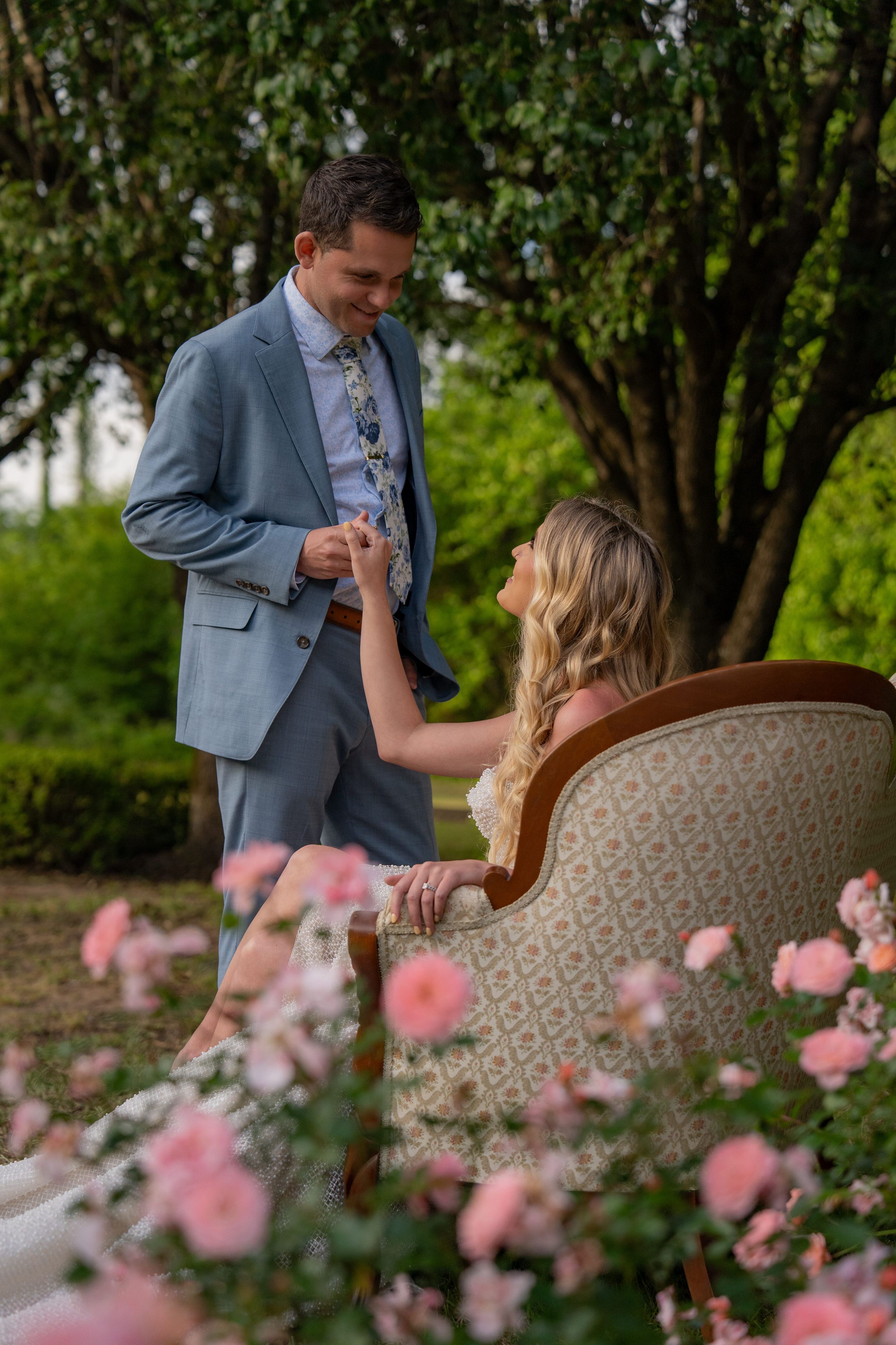 A couple shares a romantic moment on a vintage sofa outdoors with pink flowers in the foreground.