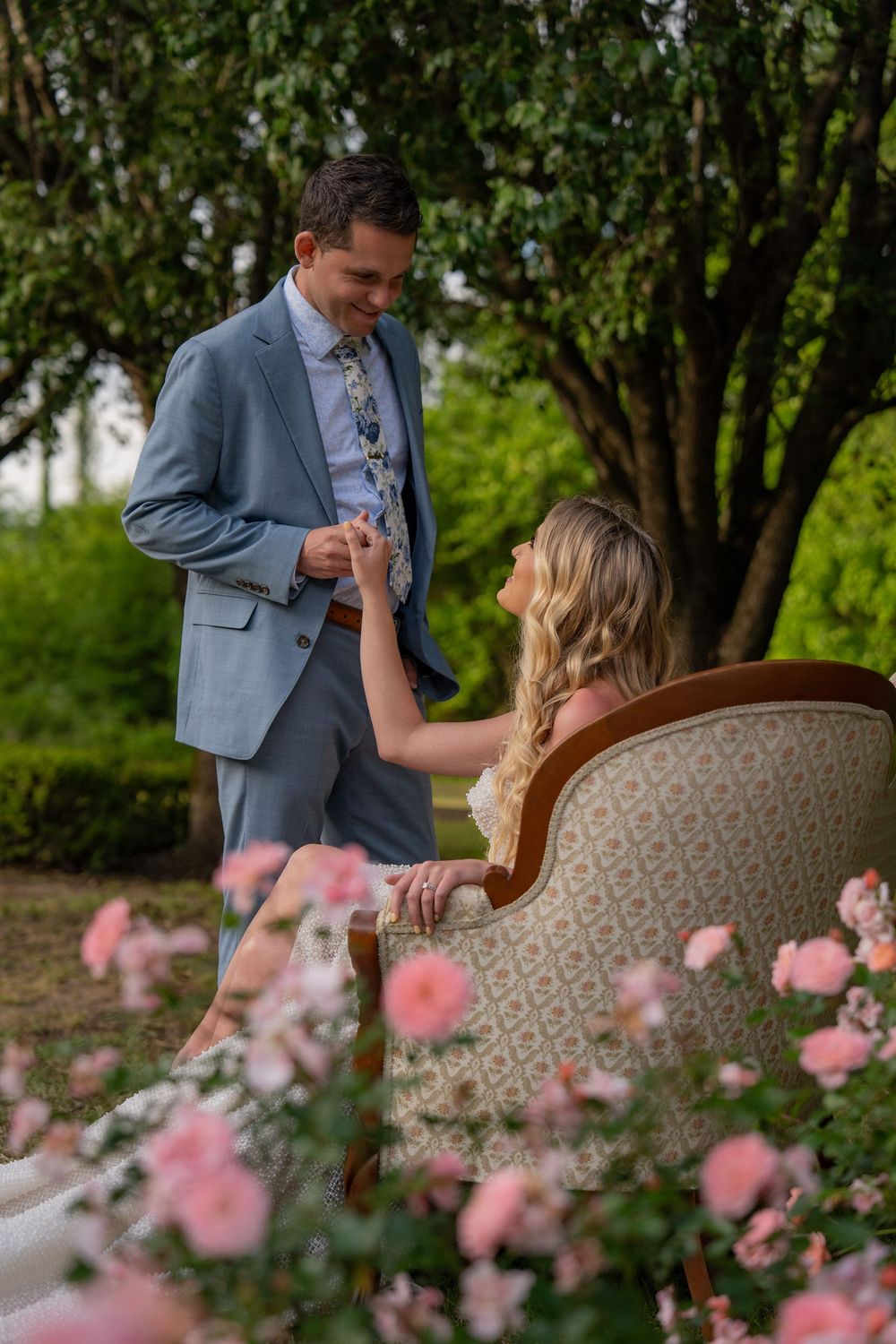 A couple shares a romantic moment on a vintage sofa outdoors with pink flowers in the foreground.