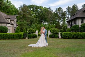 A couple embraces on a manicured lawn between two elegant houses with tall trees in the background.
