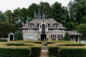 Elegant French-style mansion with manicured hedges, stone fountain, and symmetrical dormers set among tall pine trees.