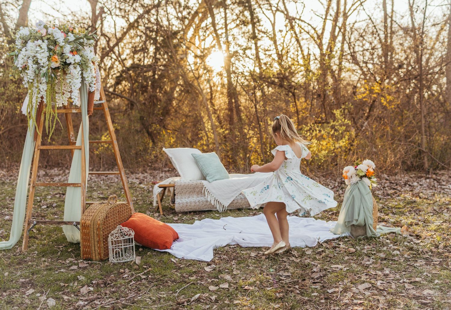 A dreamy outdoor scene with white linens, vintage furniture, and decorative elements set in a golden sunset forest setting.
