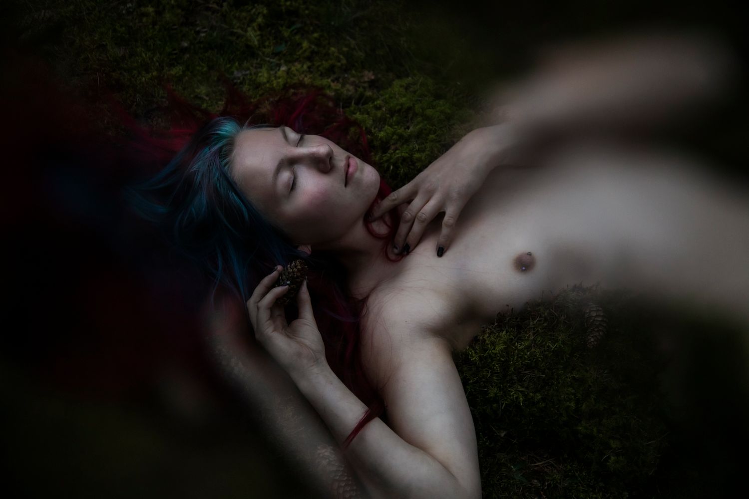 Artistic portrait of a person with blue hair lying on grass in a moody, dark setting.
