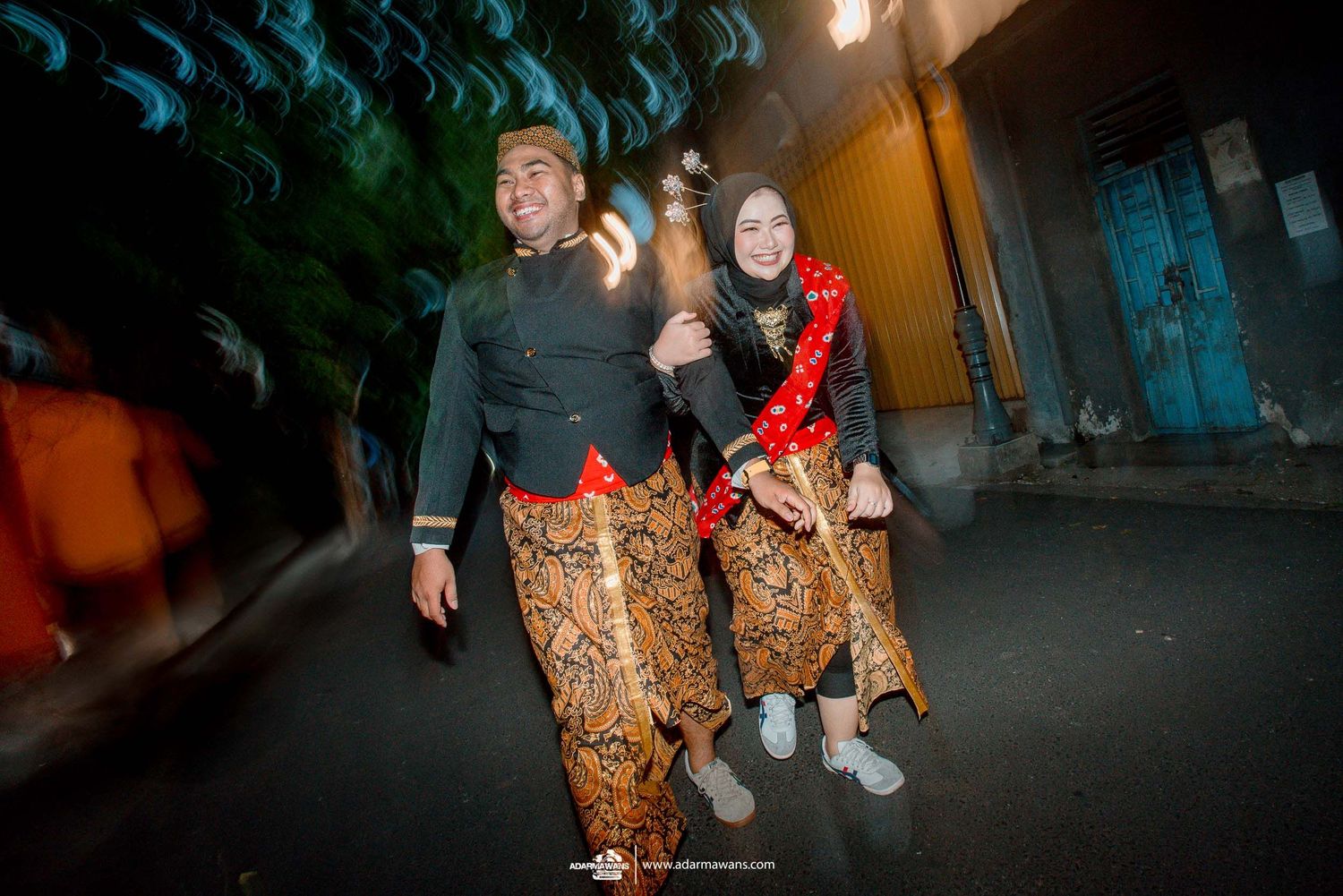 Two people in traditional Javanese batik sarongs walk together at night on a dark street.