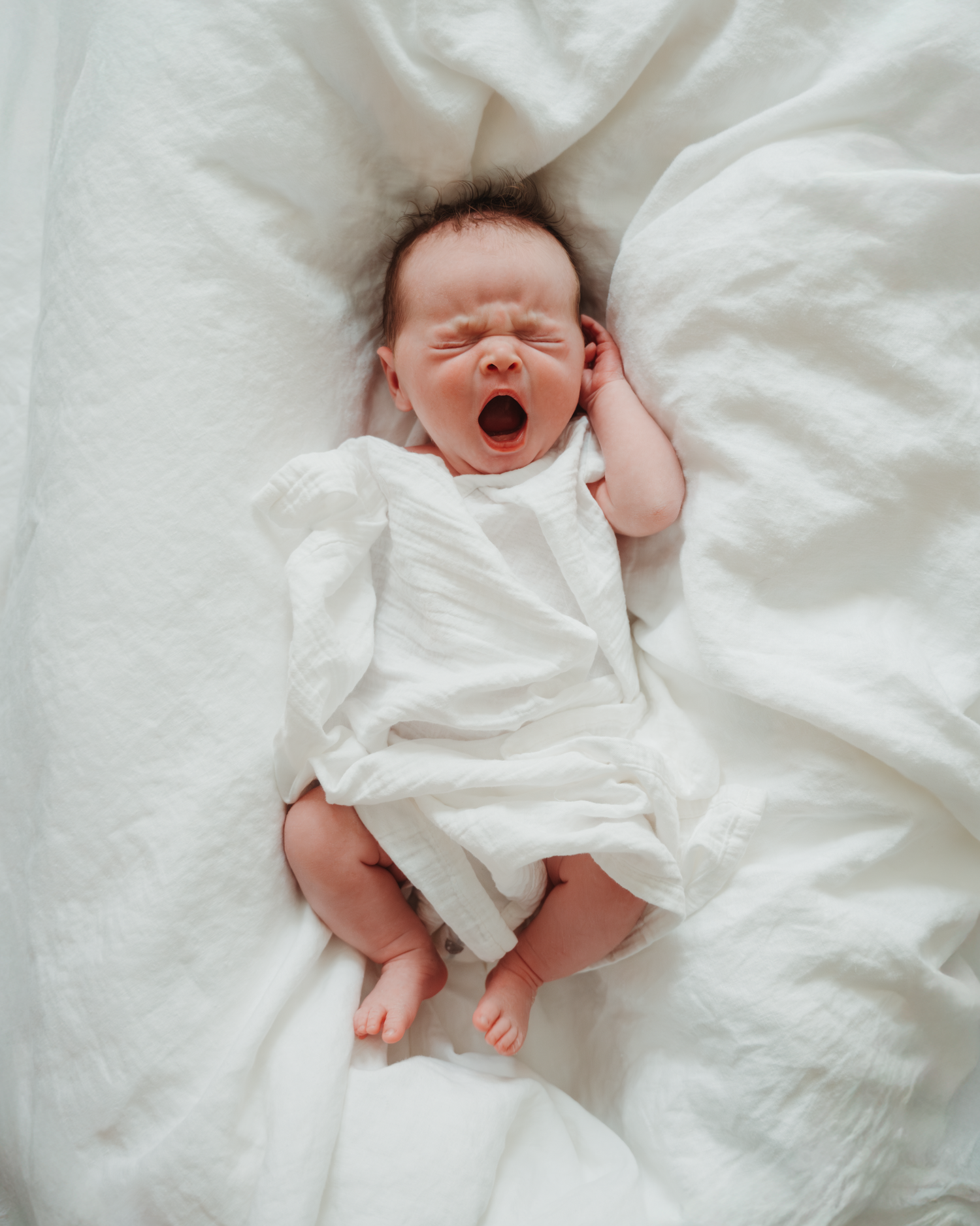 Newborn baby in white clothing yawns while lying on white bedding.