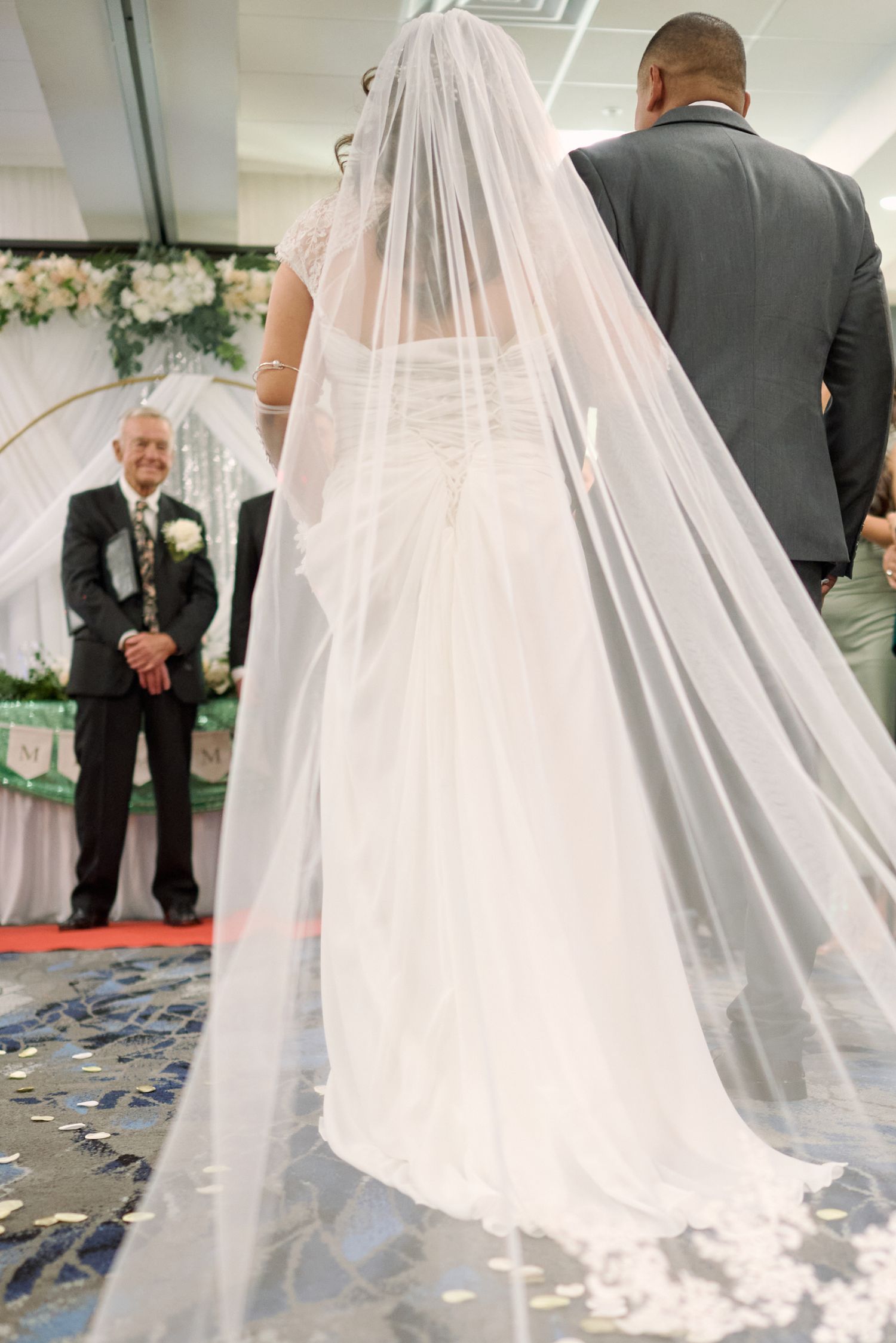 A bride in a flowing white veil and dress stands beside someone in a grey suit at a floral-decorated wedding ceremony.