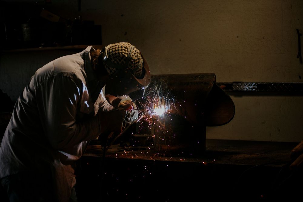 A welder in protective gear working on a metal piece in a dark workshop with bright sparks flying.