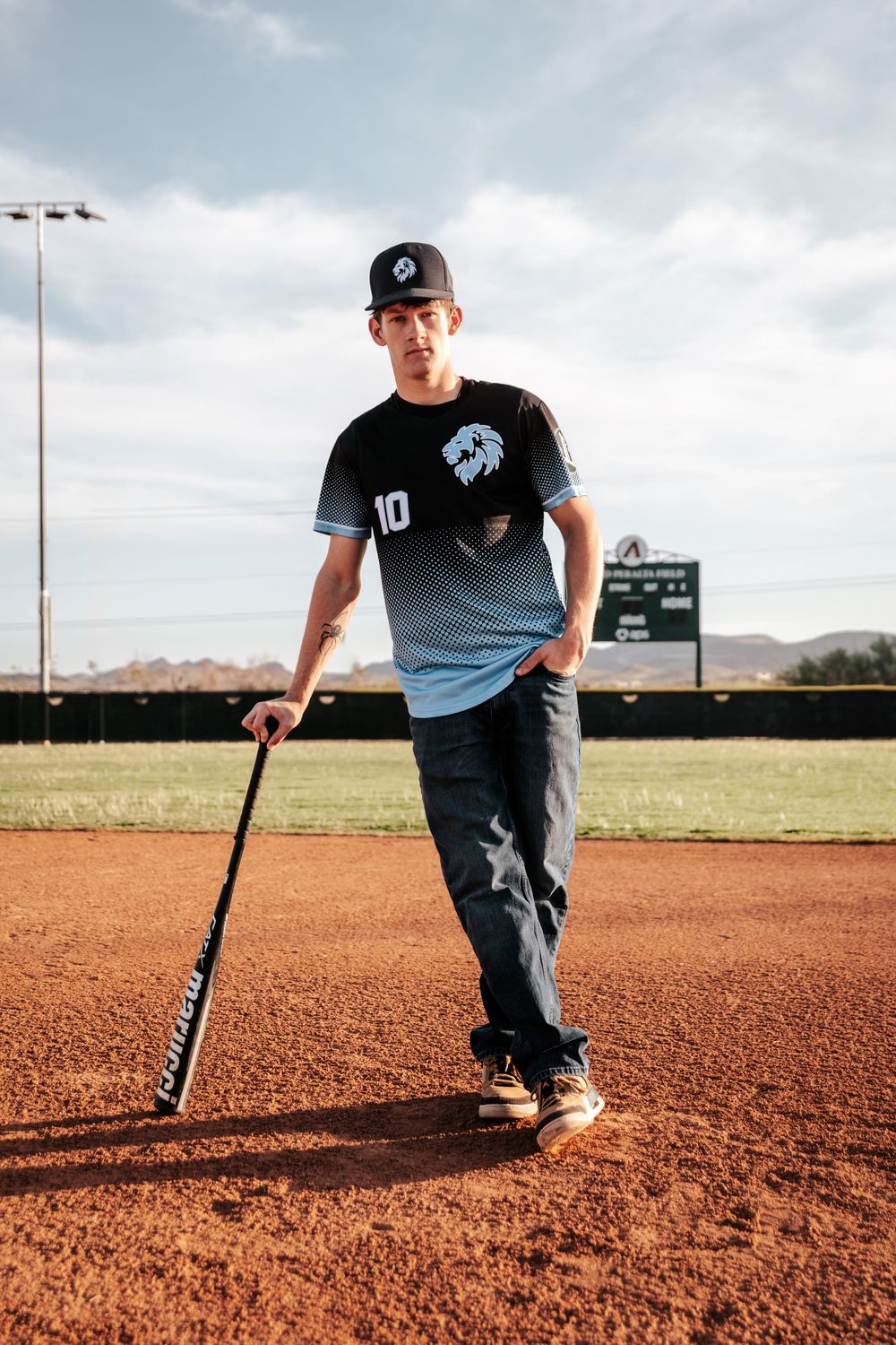 Baseball player poses with bat on dirt field with mountains and scoreboard in background.