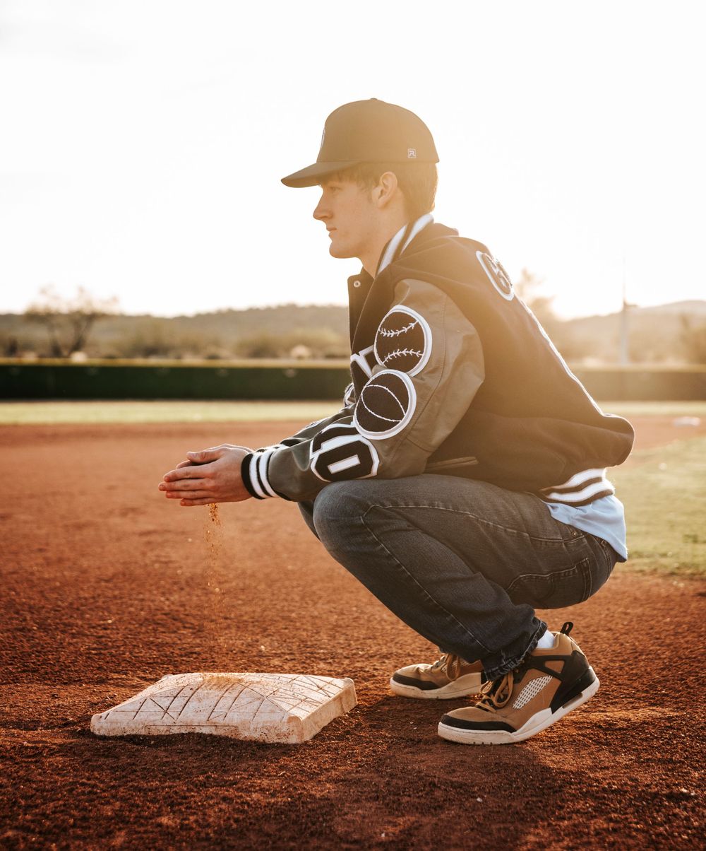 Athletic figure in varsity jacket crouches on baseball field at golden hour examining base.