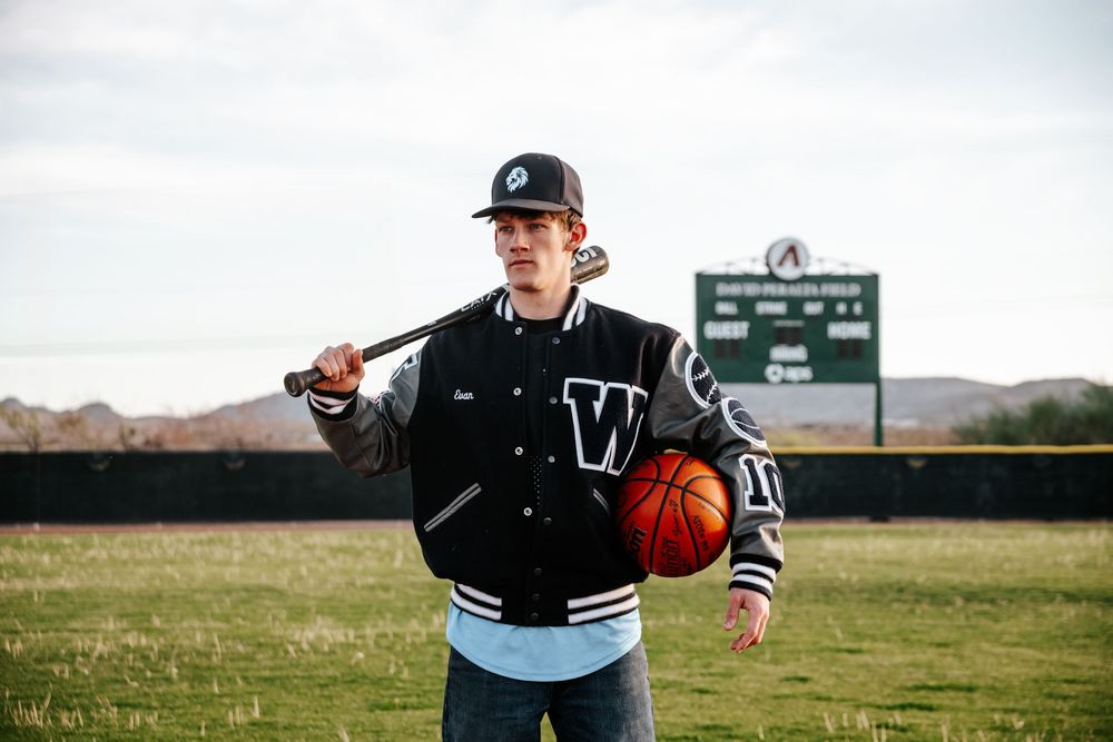 Player in black varsity jacket holds baseball bat on field with scoreboard in background.