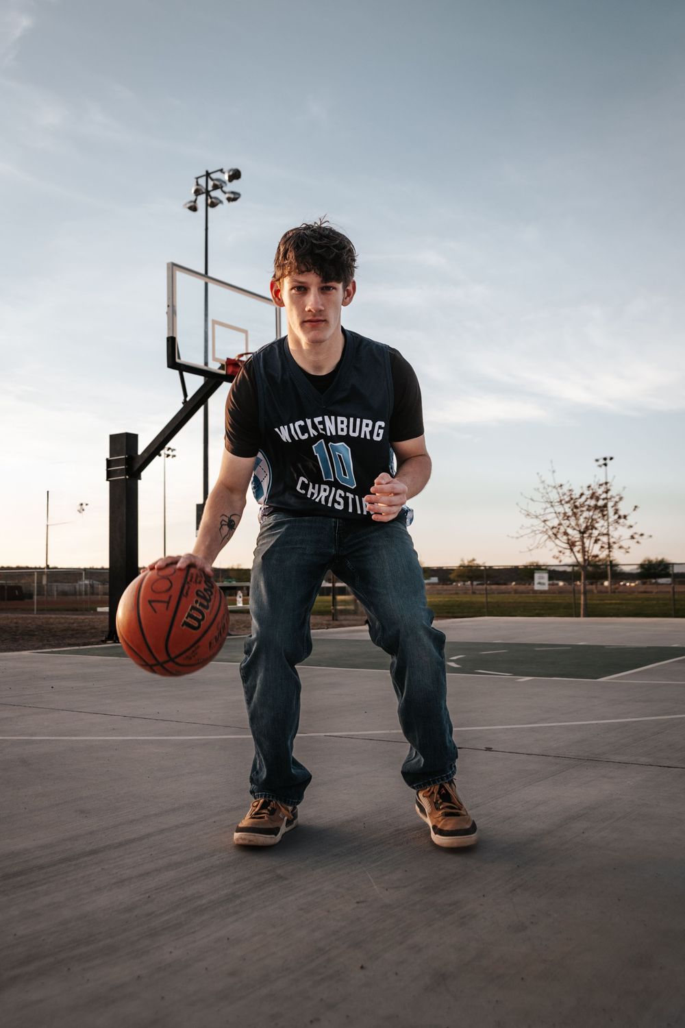 Basketball player in Wickenburg jersey dribbles on outdoor court at dusk.