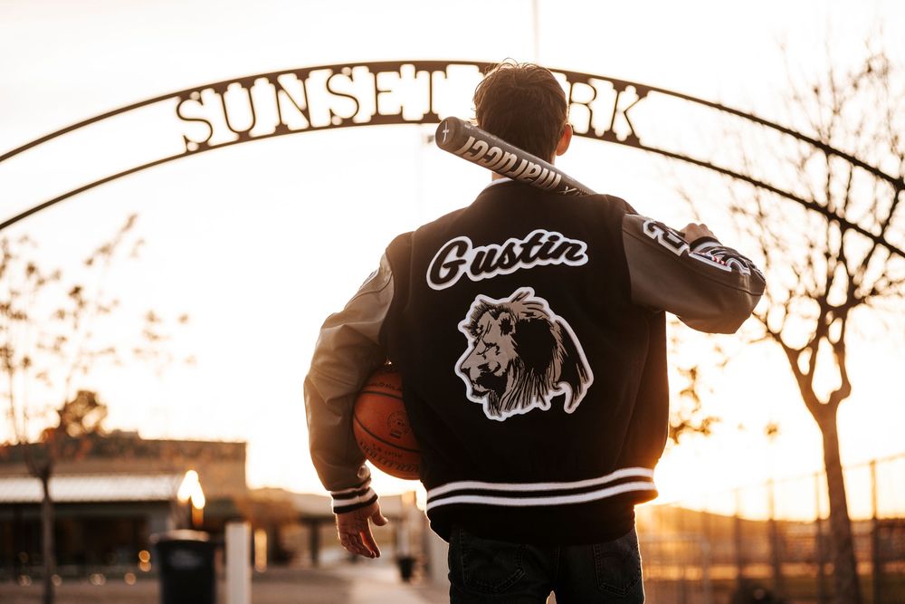 Student athlete wearing varsity jacket stands beneath Sunset Park arch at golden hour.