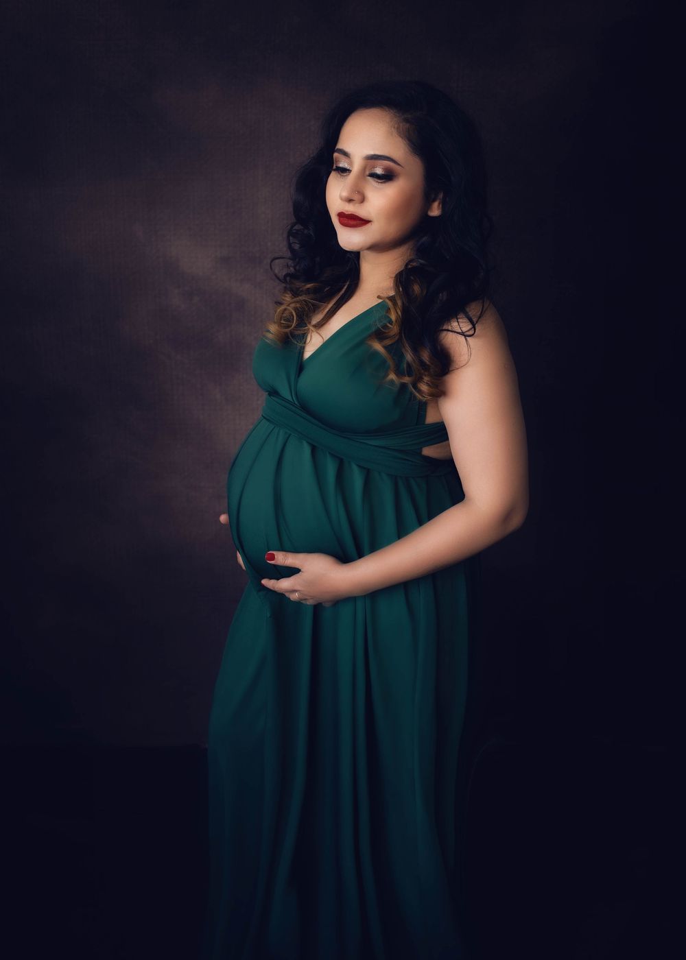 Maternity portrait of pregnant woman wearing emerald green flowing gown against dark background.