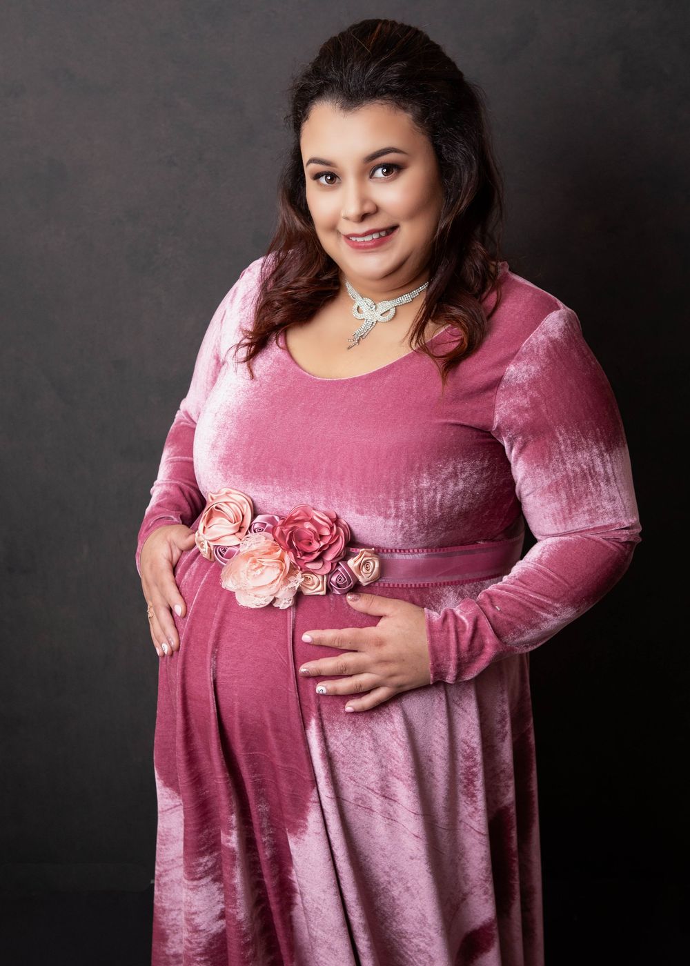 A maternity photo in a pink velvet dress with floral belt, shot against a dark gray backdrop.