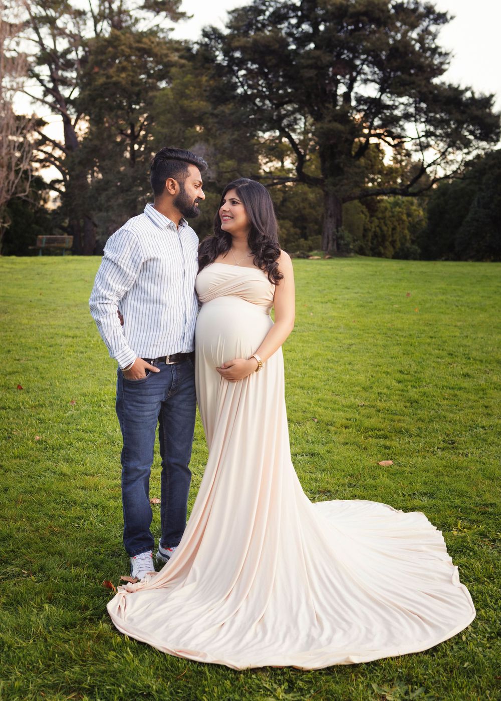 A couple in casual and formal wear share a romantic moment on a grassy field at sunset with tall trees in background.