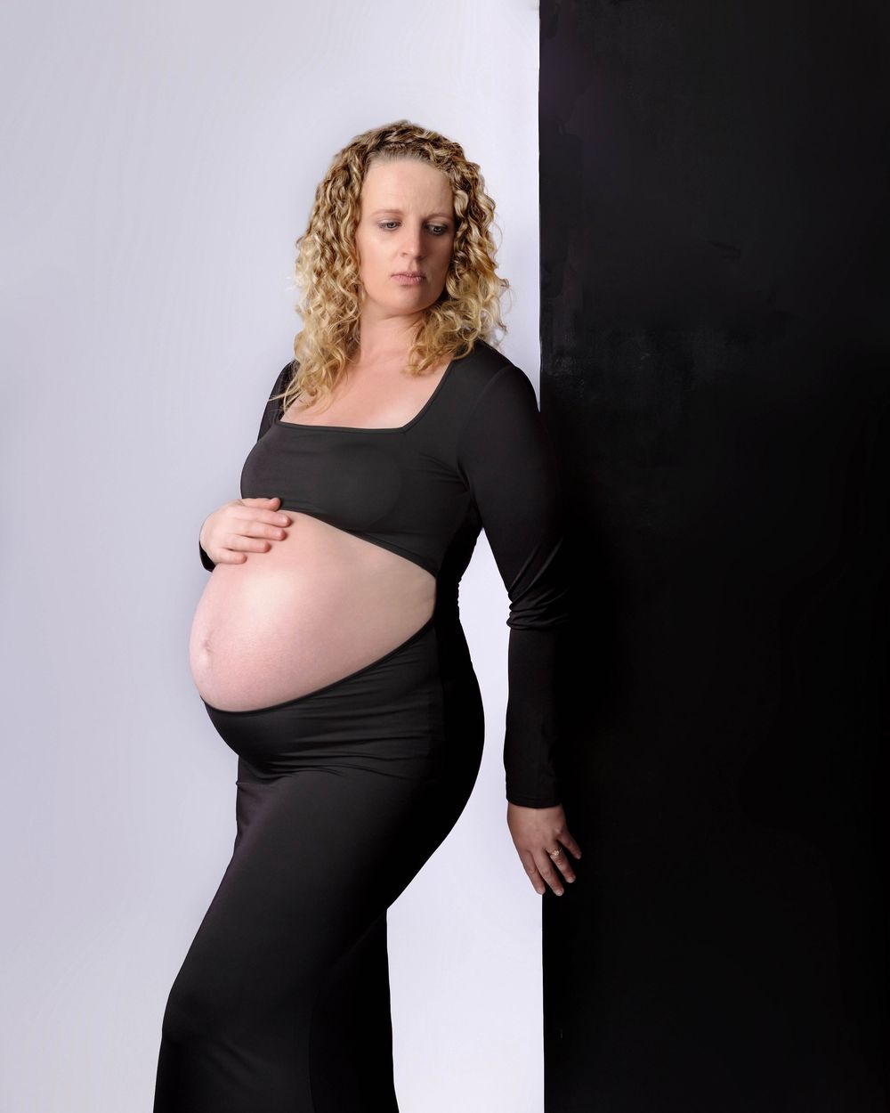 Expectant mother in black long-sleeved dress poses against contrasting light and dark background in maternity photo.