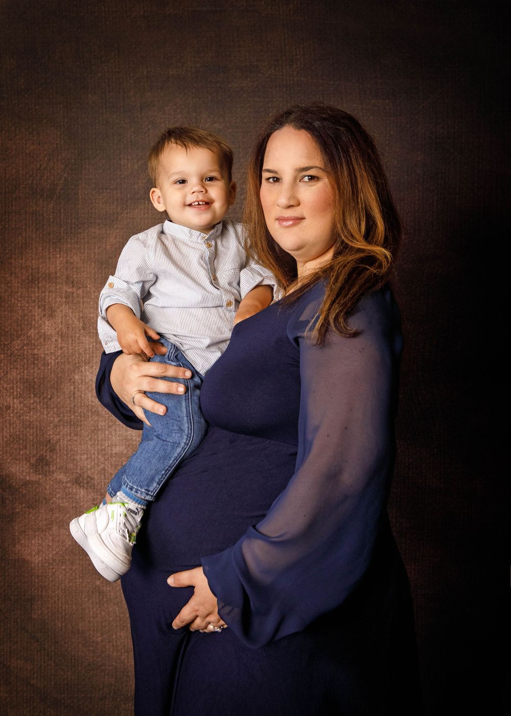 A pregnant woman in a navy blue dress poses with a toddler wearing white and jeans against a dark studio backdrop.