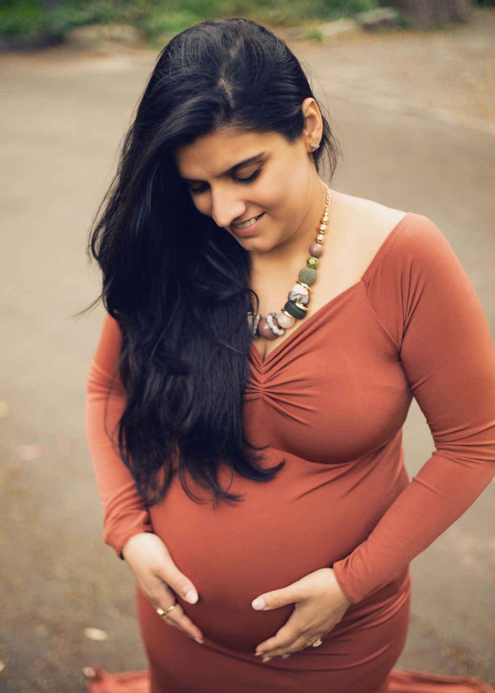 A pregnant woman in a terracotta-colored dress touches her belly while looking down with flowing dark hair.