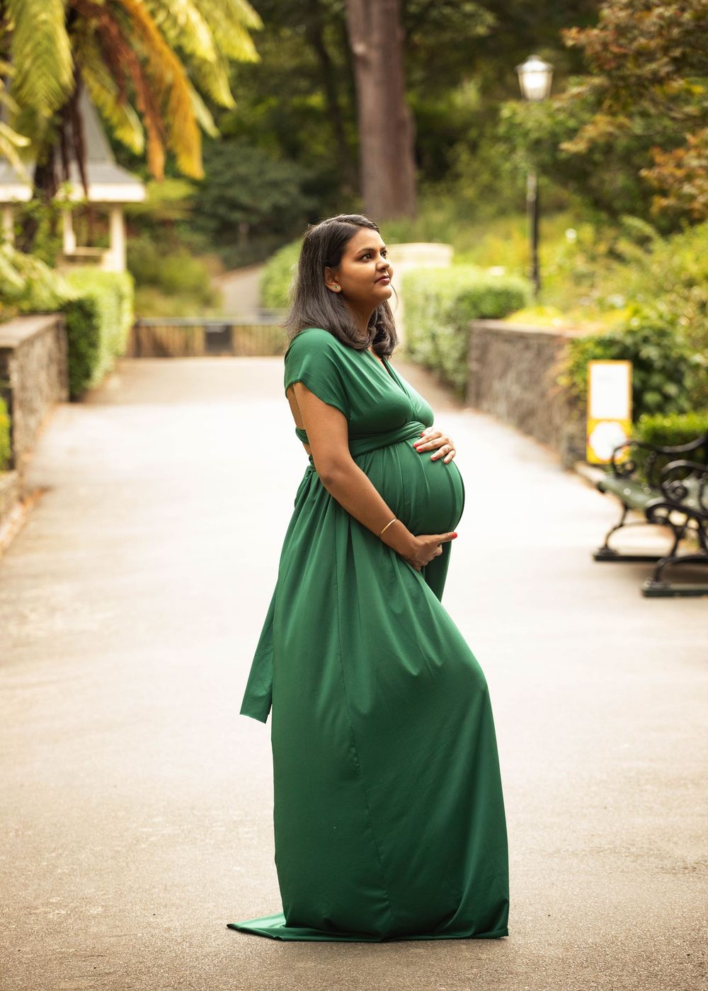 Expectant person wearing a flowing emerald green maxi dress poses thoughtfully on a tree-lined path with benches.
