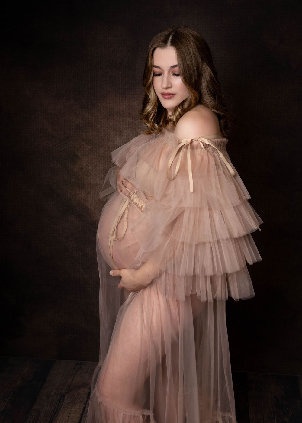 Artistic maternity portrait in blush pink tulle dress with tiered ruffles against dark moody backdrop.