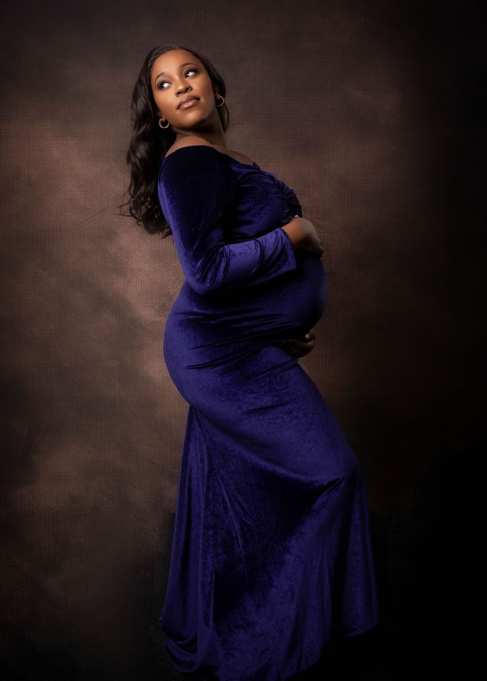 Beautiful maternity portrait of a woman in a deep purple velvet gown posing against a dark brown backdrop.