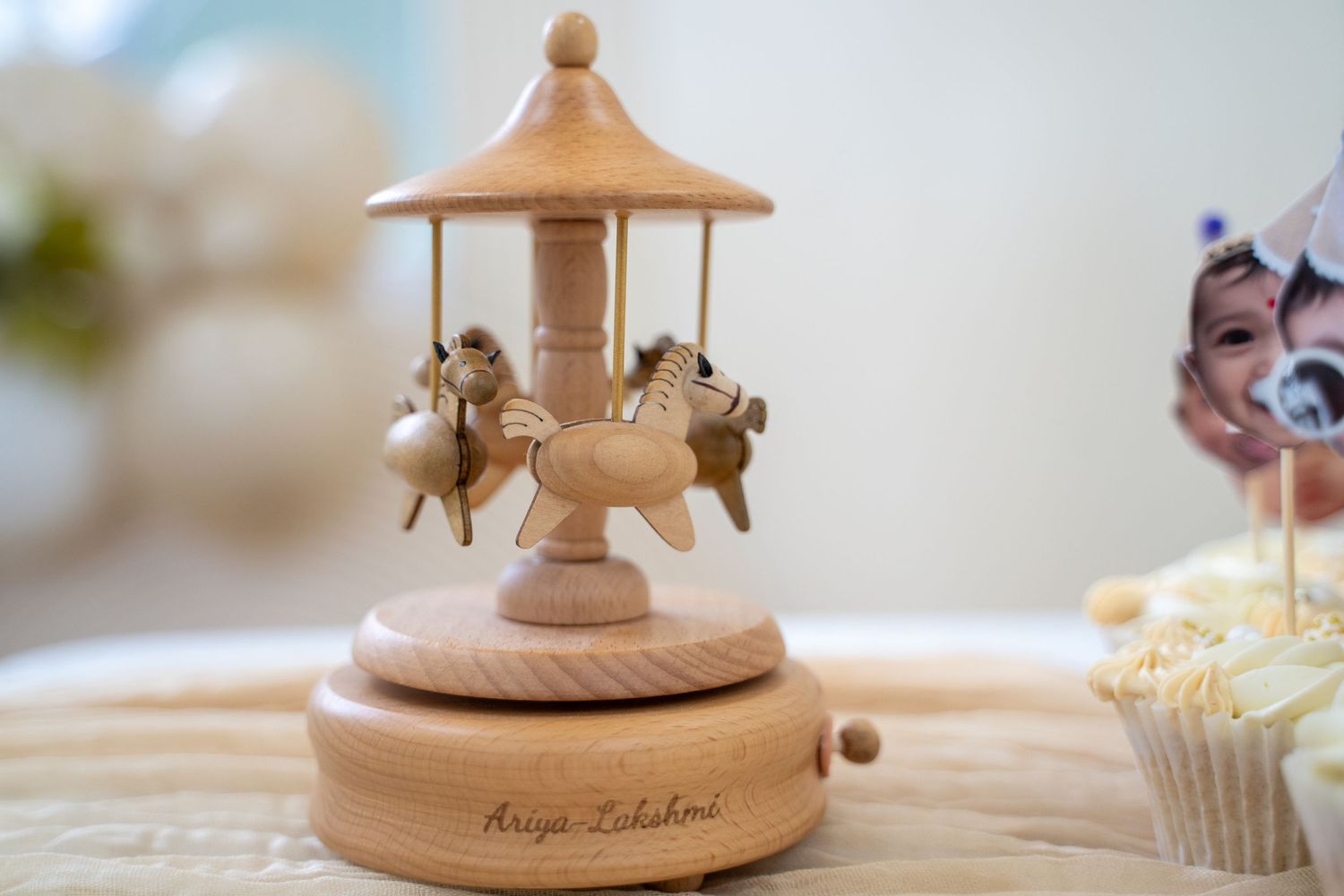 A wooden carousel music box with angels or cherubs mounted on a light colored pedestal sits next to blurred cupcakes.