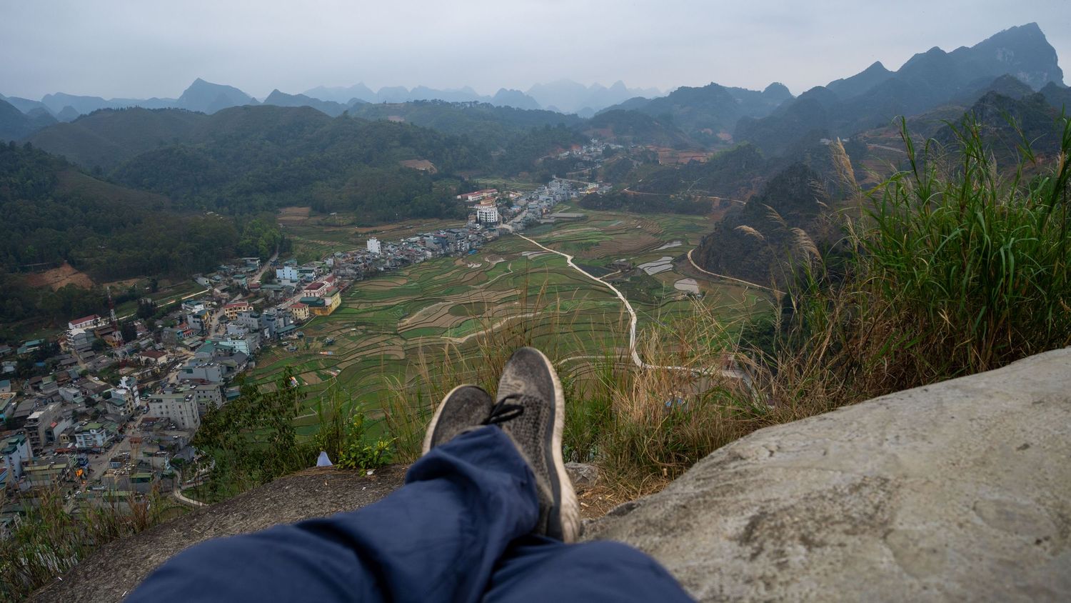 First-person view of legs dangling from a mountain viewpoint overlooking a valley town.