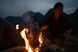 Campfire flames illuminate a nighttime gathering in the wilderness with mountains in background.