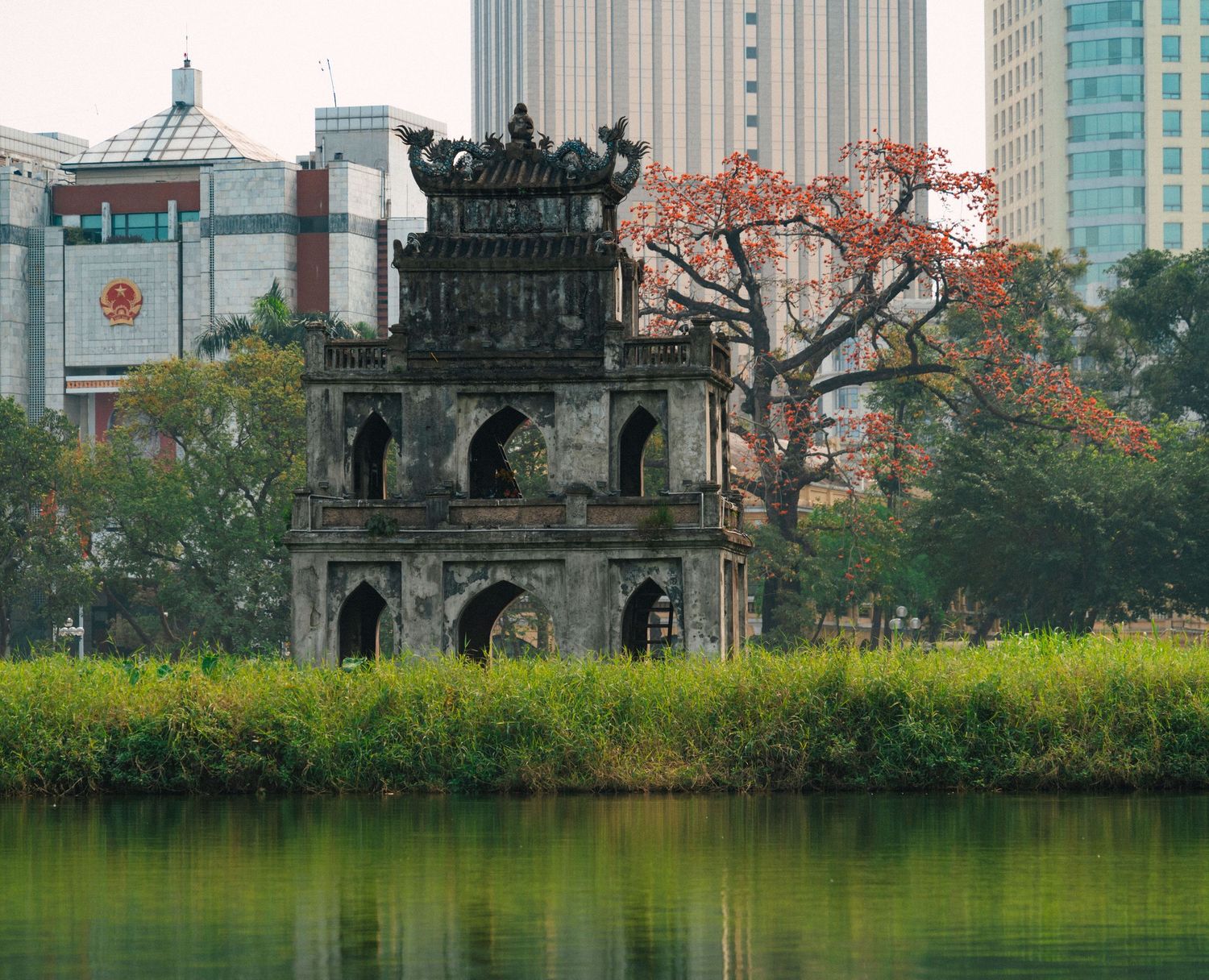Traditional Asian architecture along a tranquil lake with autumn trees and modern buildings in background.