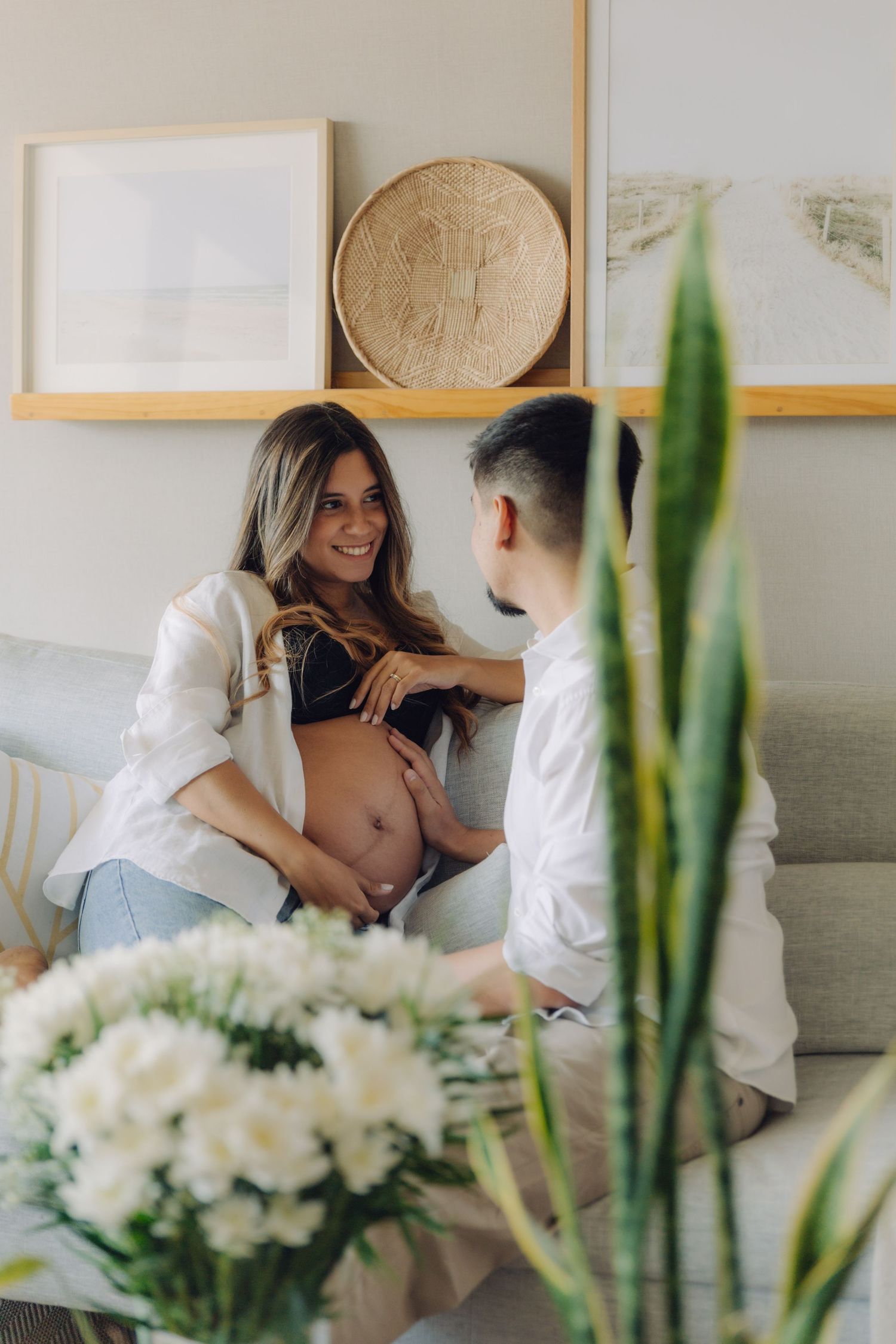 Smiling pregnant woman and partner tenderly touching baby bump on couch surrounded by flowers.