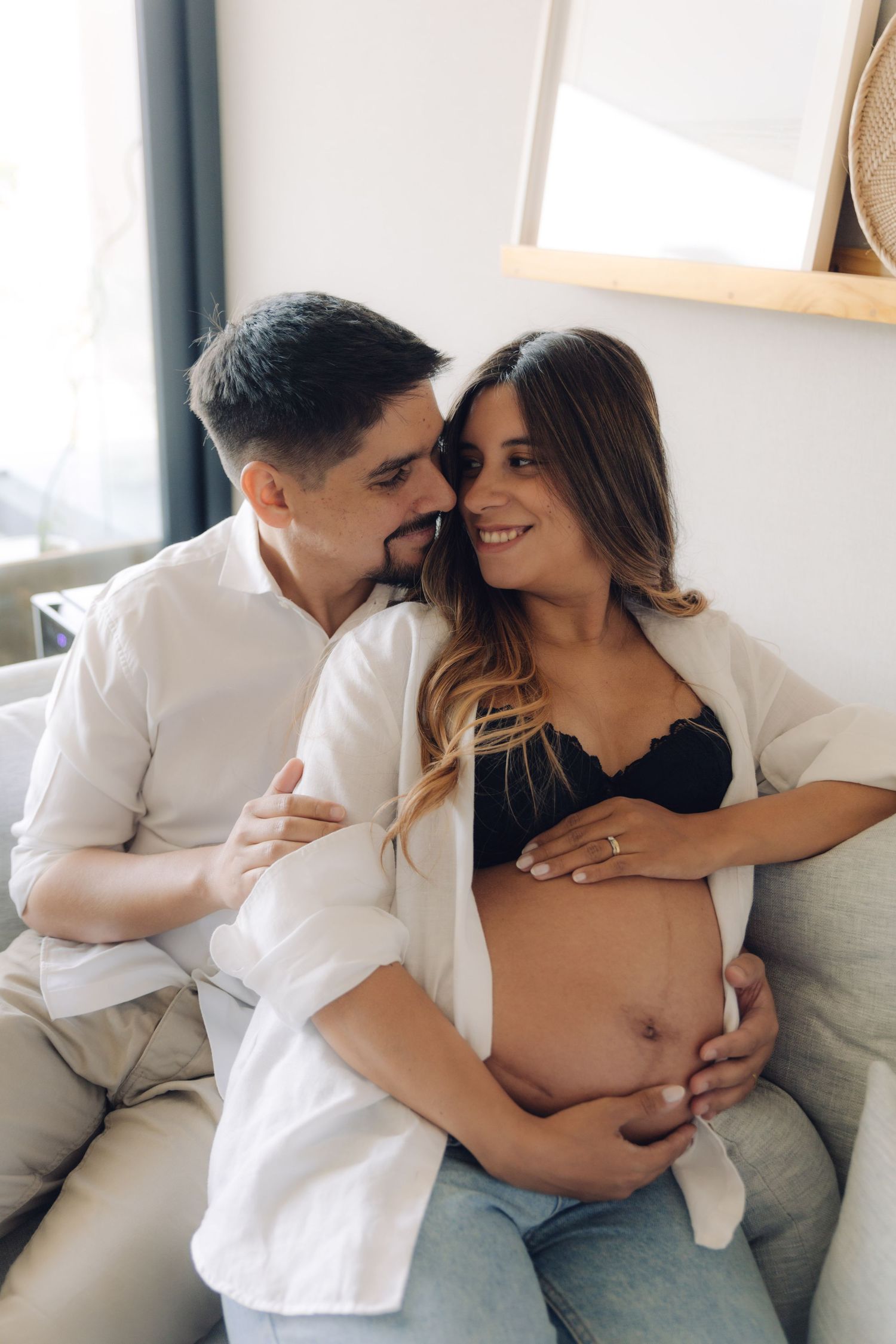 Couple sitting on couch at home, man embracing smiling pregnant woman showing her bare belly.
