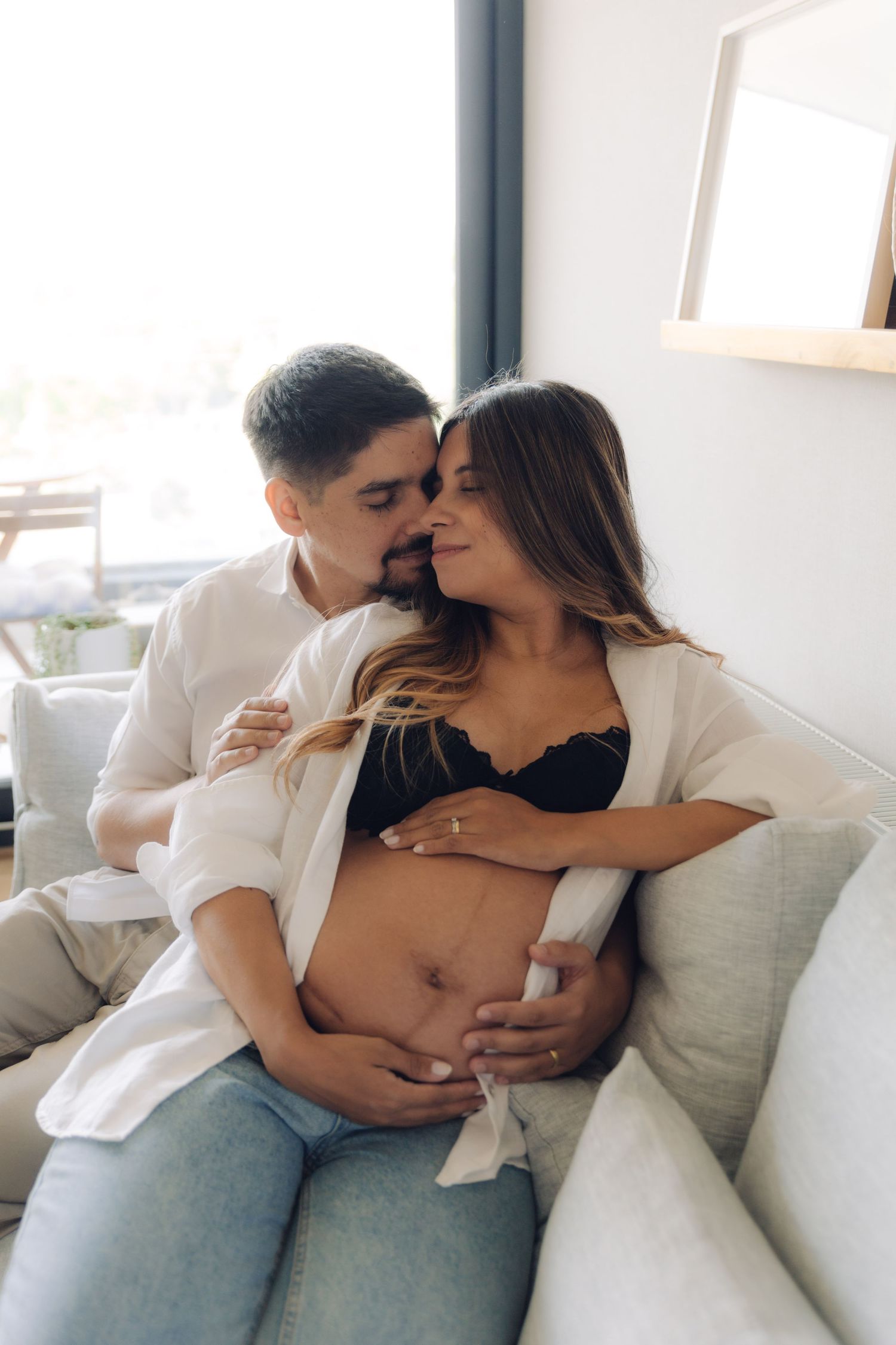 Loving couple embracing on sofa, man kissing pregnant woman's temple while cradling her baby bump.
