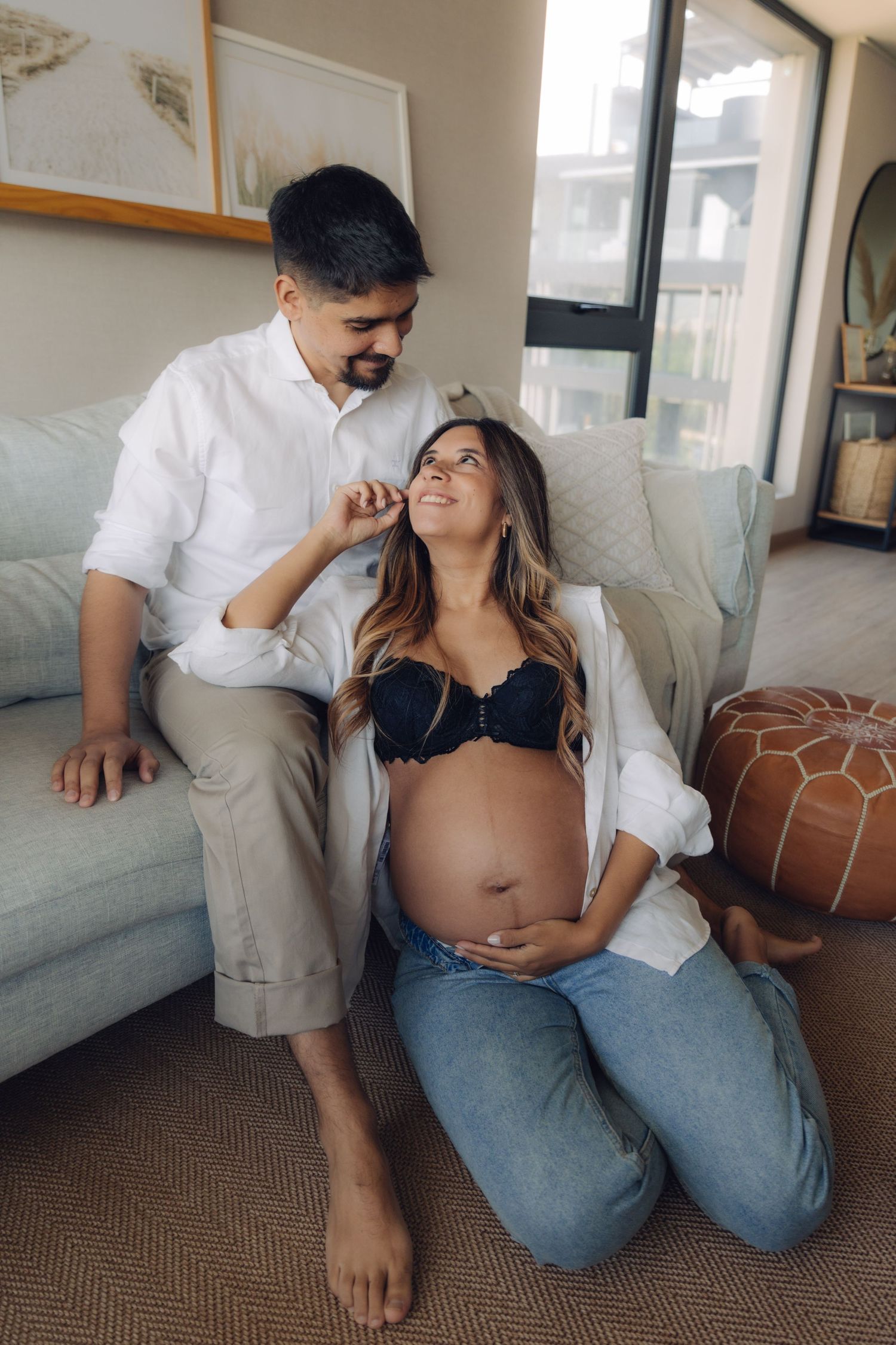Pregnant woman sitting on floor looking up lovingly at partner seated on couch behind her.