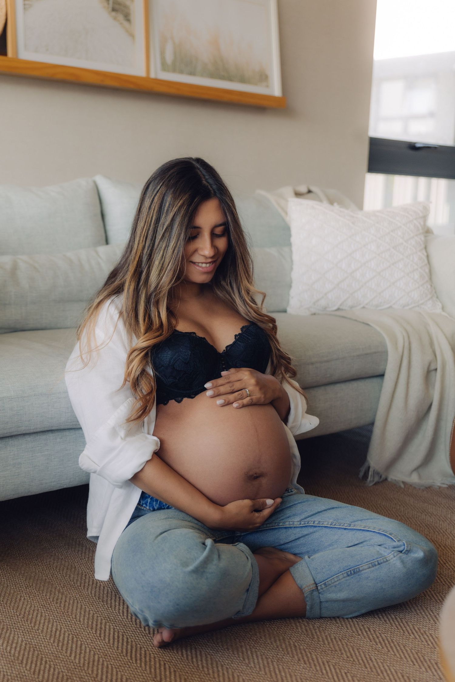Happy pregnant woman sitting cross-legged on floor cradling her bare baby bump in black bra.