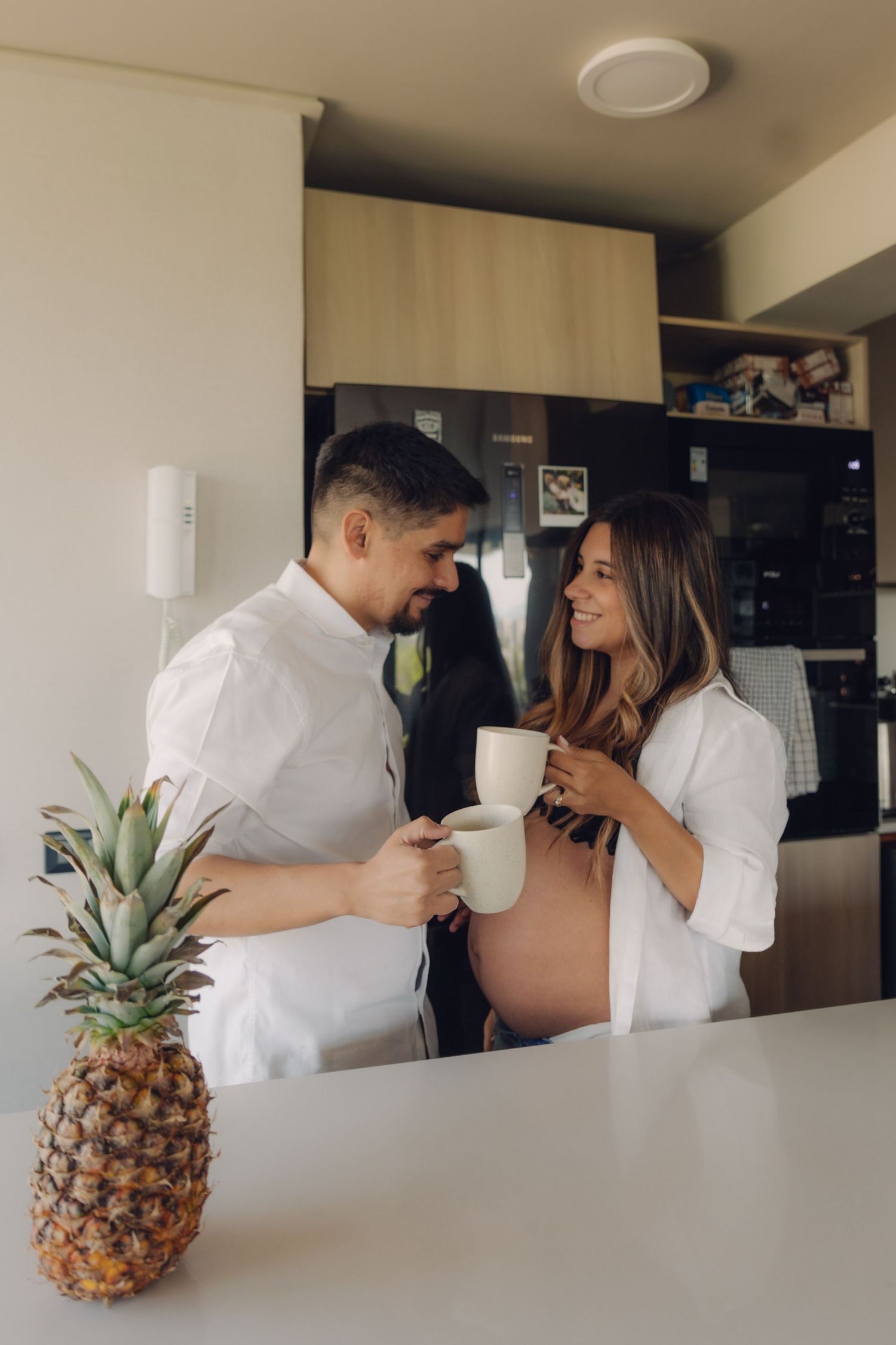 Smiling pregnant couple toasting mugs in modern kitchen with pineapple on counter.