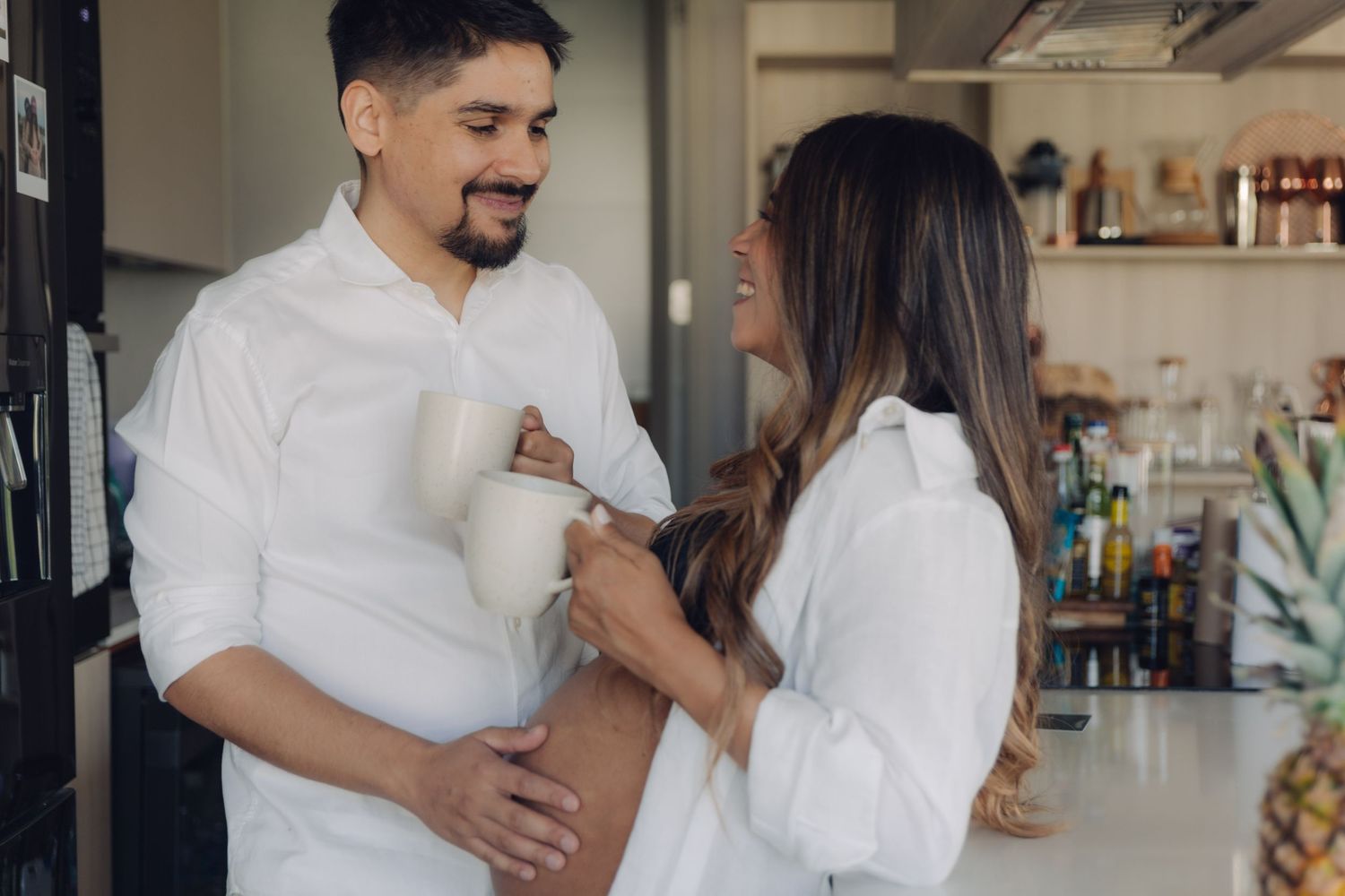 Couple in matching white shirts sharing a warm moment in kitchen, man holding pregnant belly.
