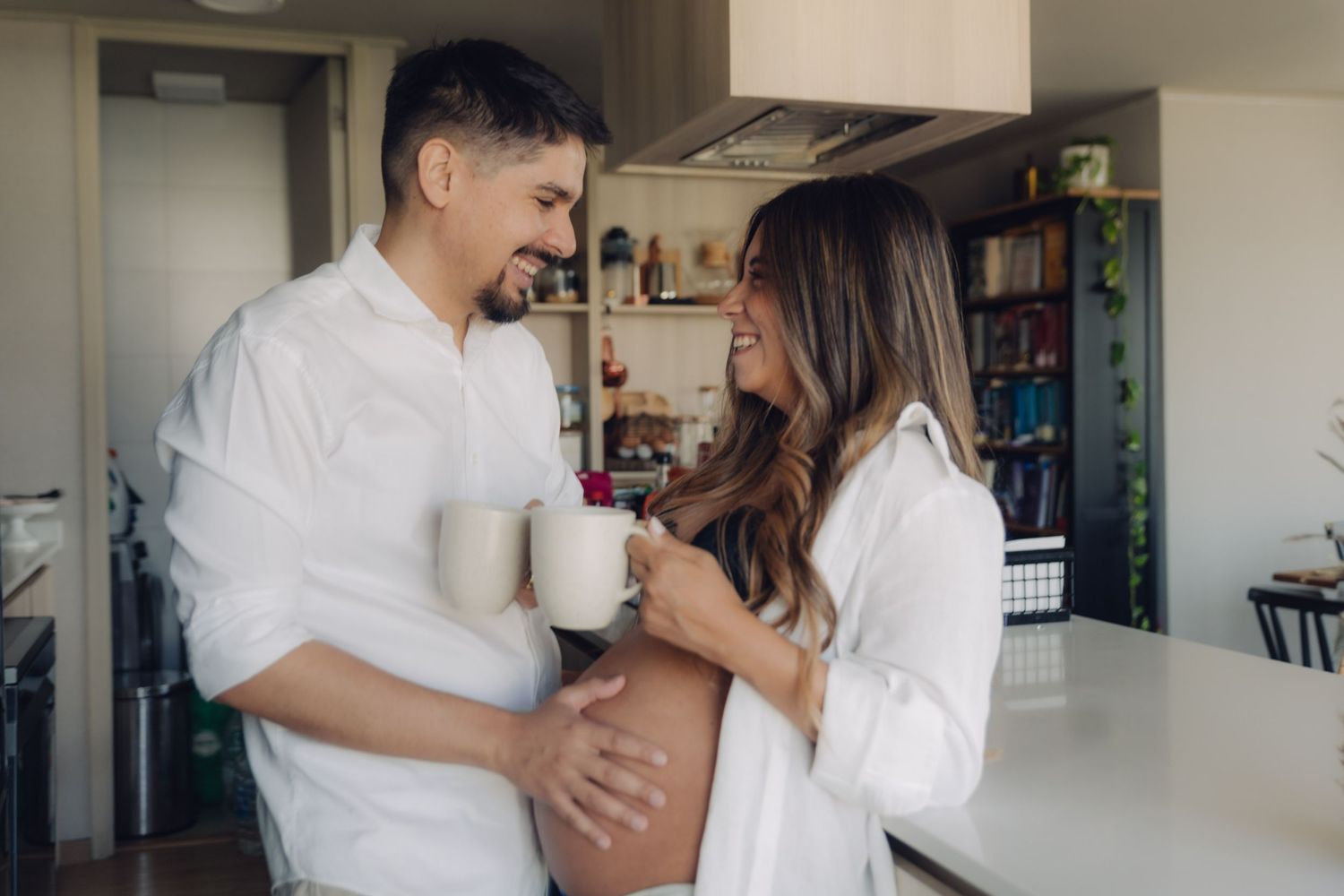 Happy expecting couple in white shirts clinking mugs while man holds pregnant woman's belly.