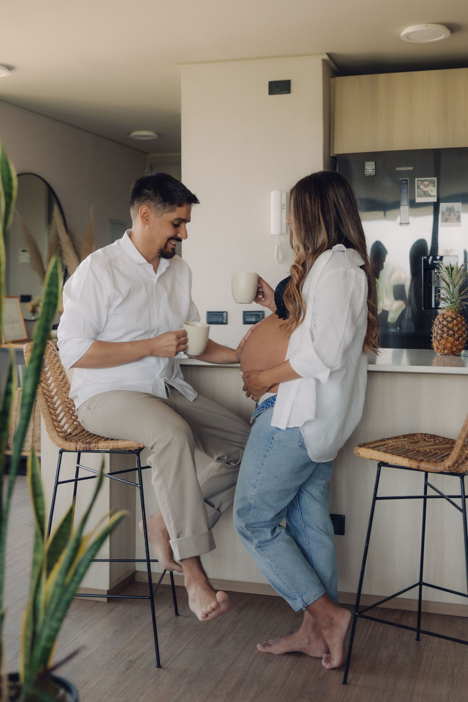 Pregnant couple sharing a tender moment in a modern kitchen, man seated on rattan stool holding coffee cup.