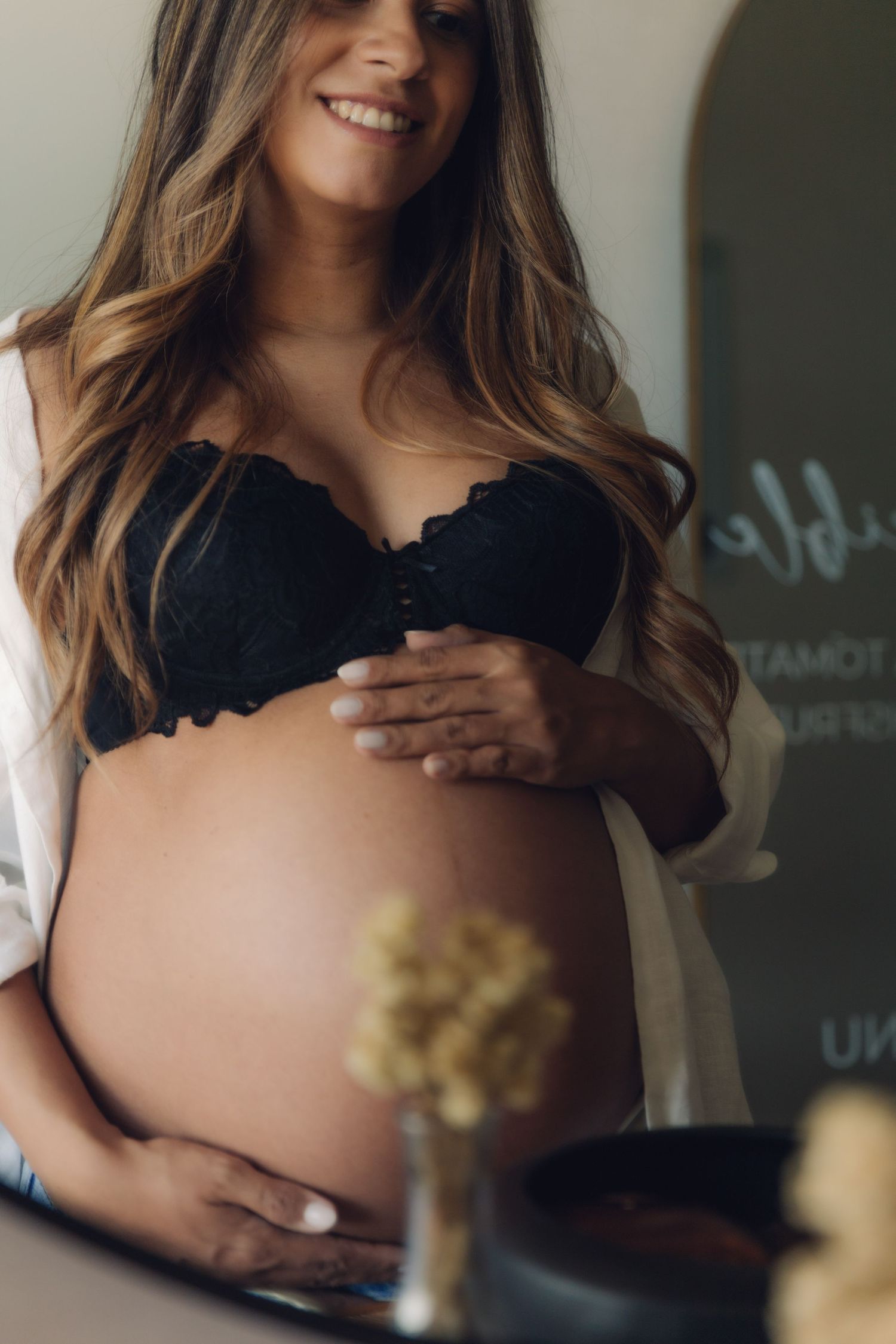 Smiling pregnant woman in black lace bra and open white shirt cradling her bare baby bump.